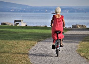 eldre dame på sykkel, bildet er tatt bakfra, hun har rød kjole på, himmel og hav i bakgrunnen
